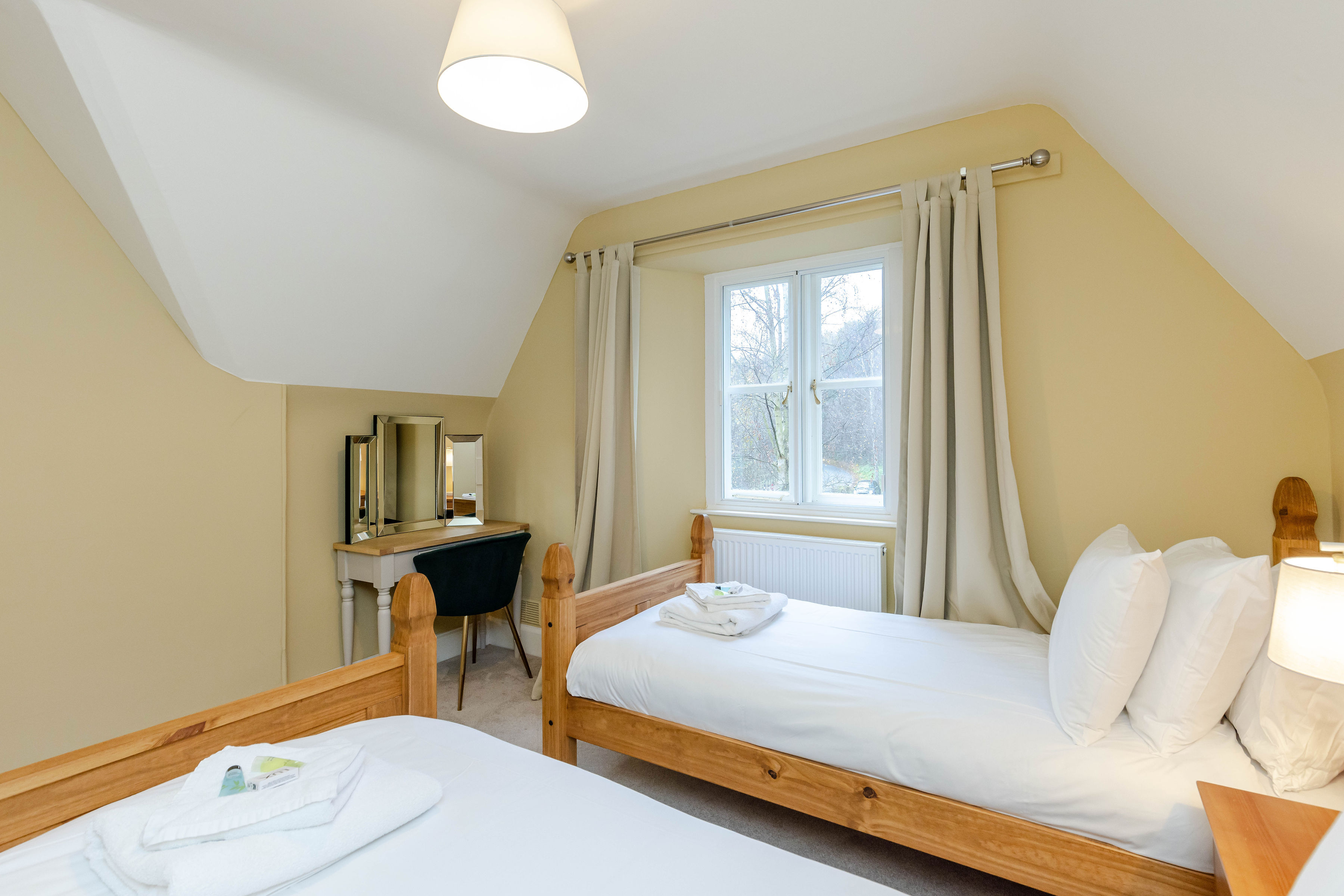 A bedroom with soft yellow walls, two wooden twin beds and a view of trees from the window