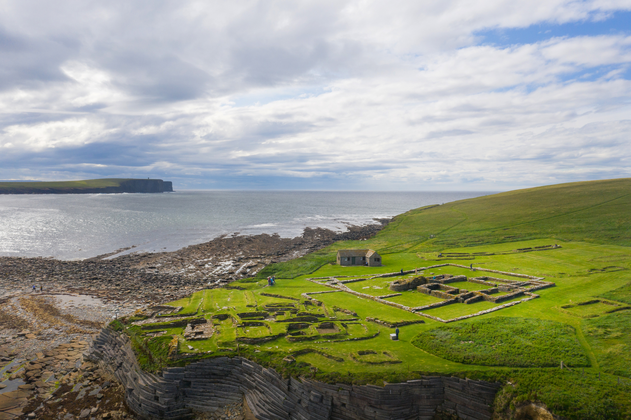 Aerial view of the Brough of Birsay showing green fields and the outlines of archaeological ruins on a clifftop overlooking the sea.