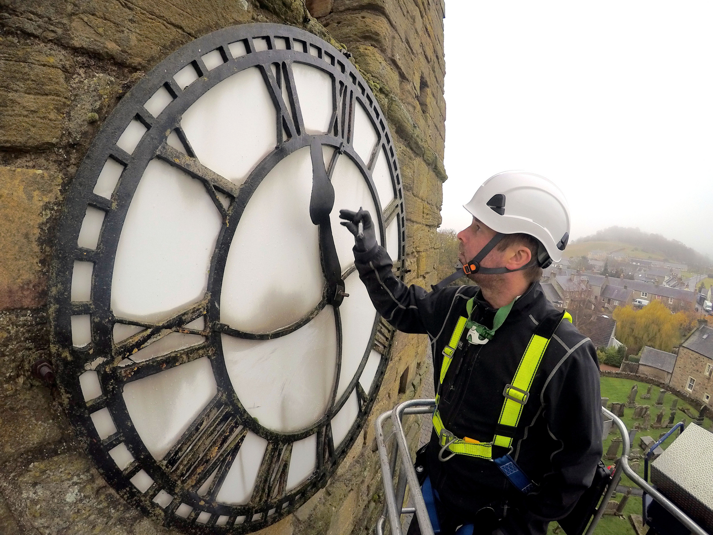 A clock technician wearing safety gear works on a large Roman‑numeral clock face of a stone church tower.