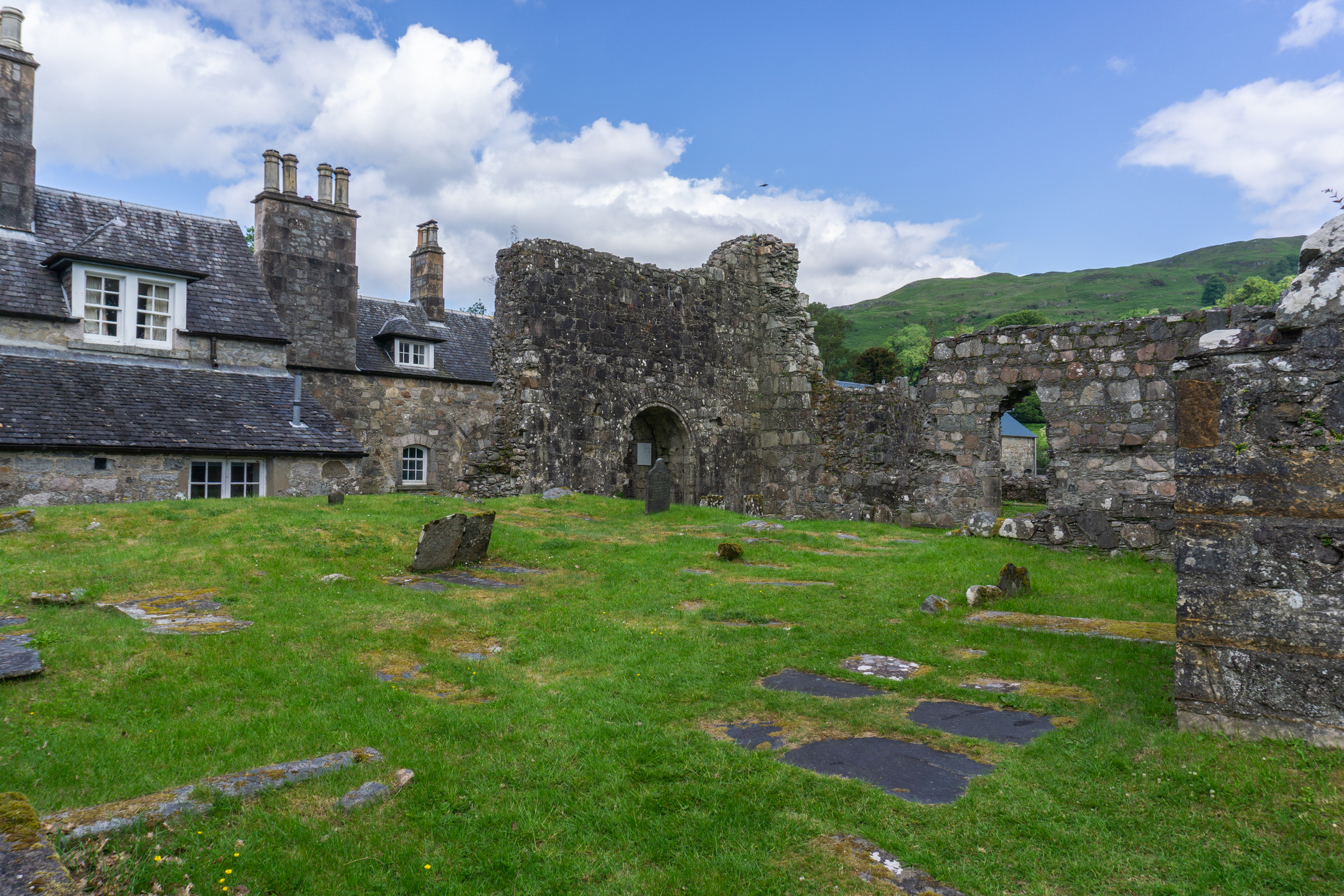 A wide view of Ardchattan Priory’s stone ruins beside an adjoining historic house with slate roofs and chimneys, surrounded by grass and remnants of old grave markers, with green hills rising behind.