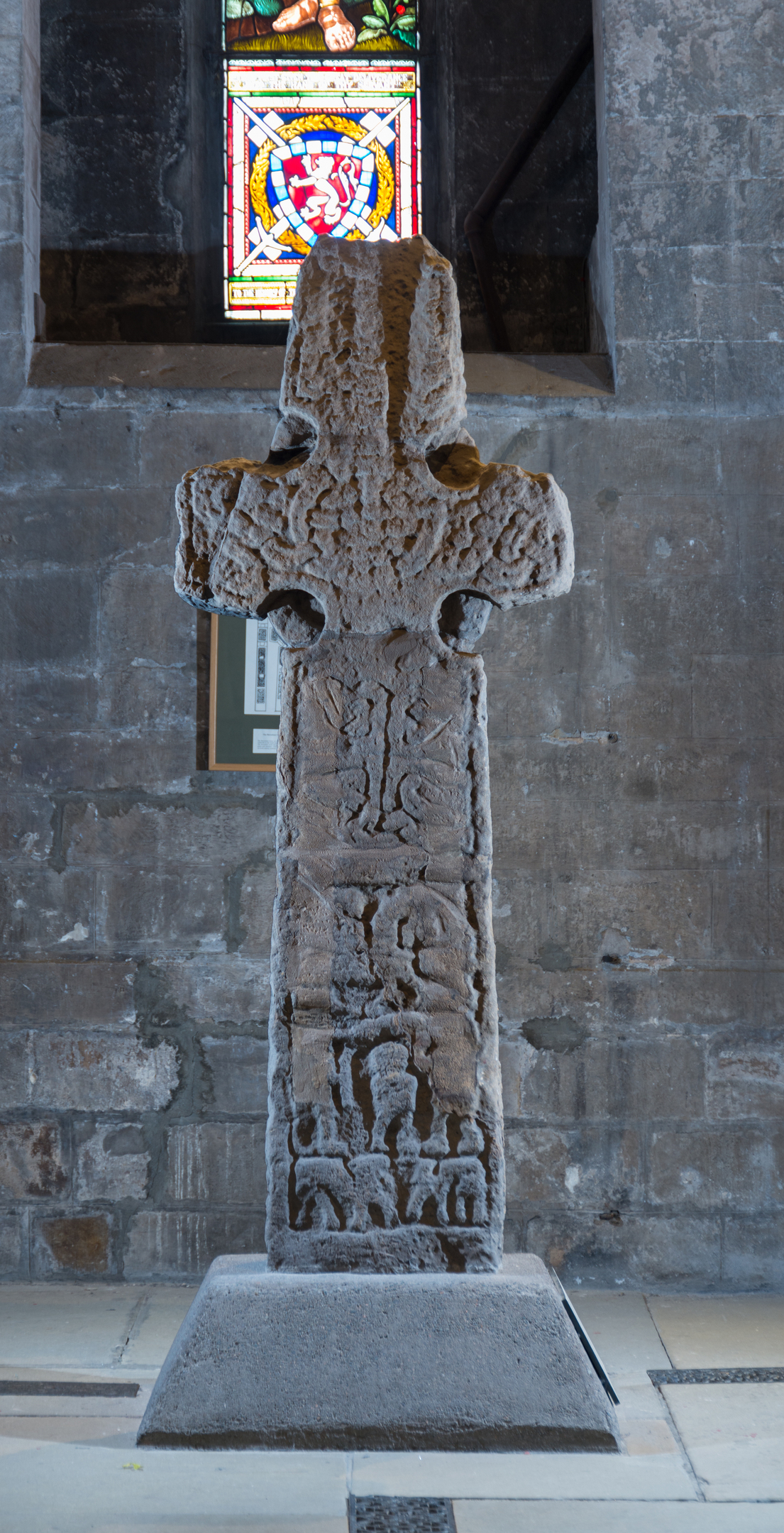 Carved stone cross with interior wall of a church or abbey behind it with part of a stained glass window