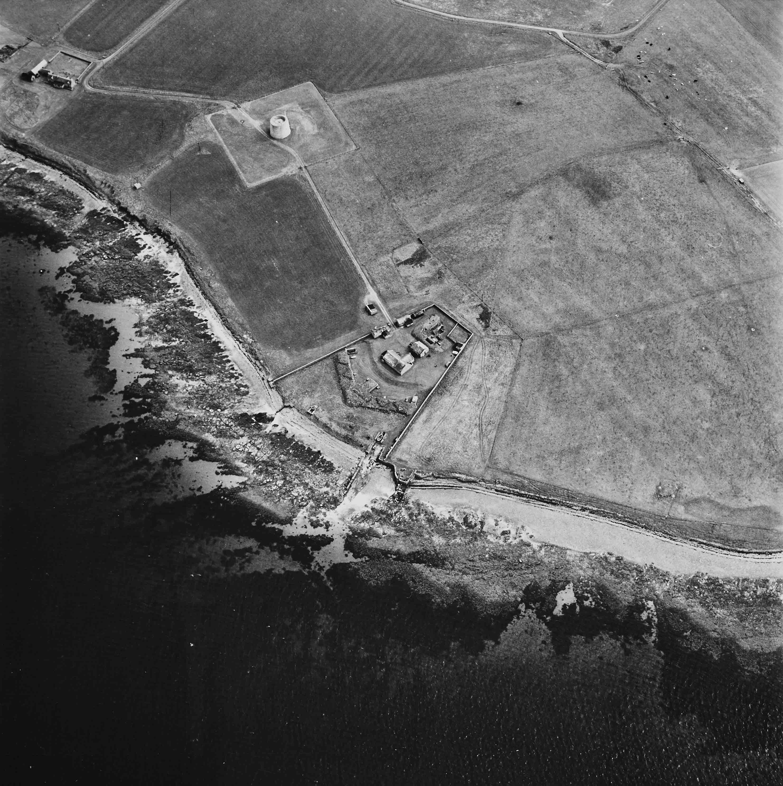 An aerial view of a tower and barracks on the edge of the sea, surrounded by grassy fields