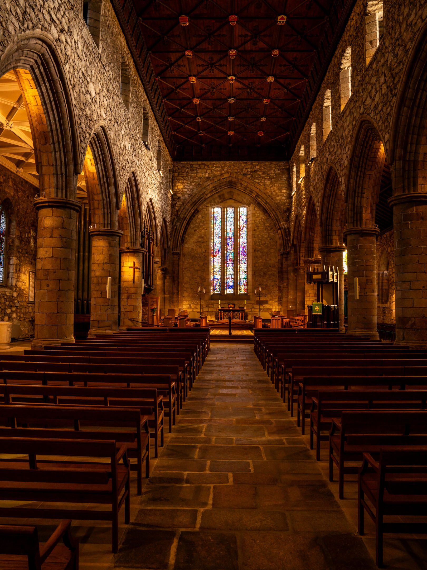 Nave interior with rows of wooden pews, stone arches and stained-glass above altar.