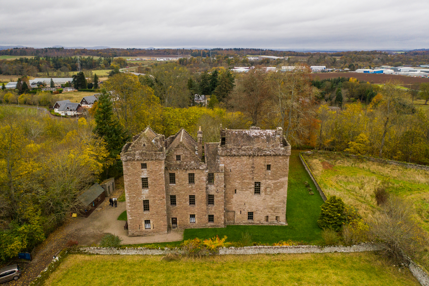 Aerial view of a stone historic castle. There are multiple large windows on the different components of the castle, and it has a darker roof which stands out. There are walls and fences around it, and there are trees in the background. 