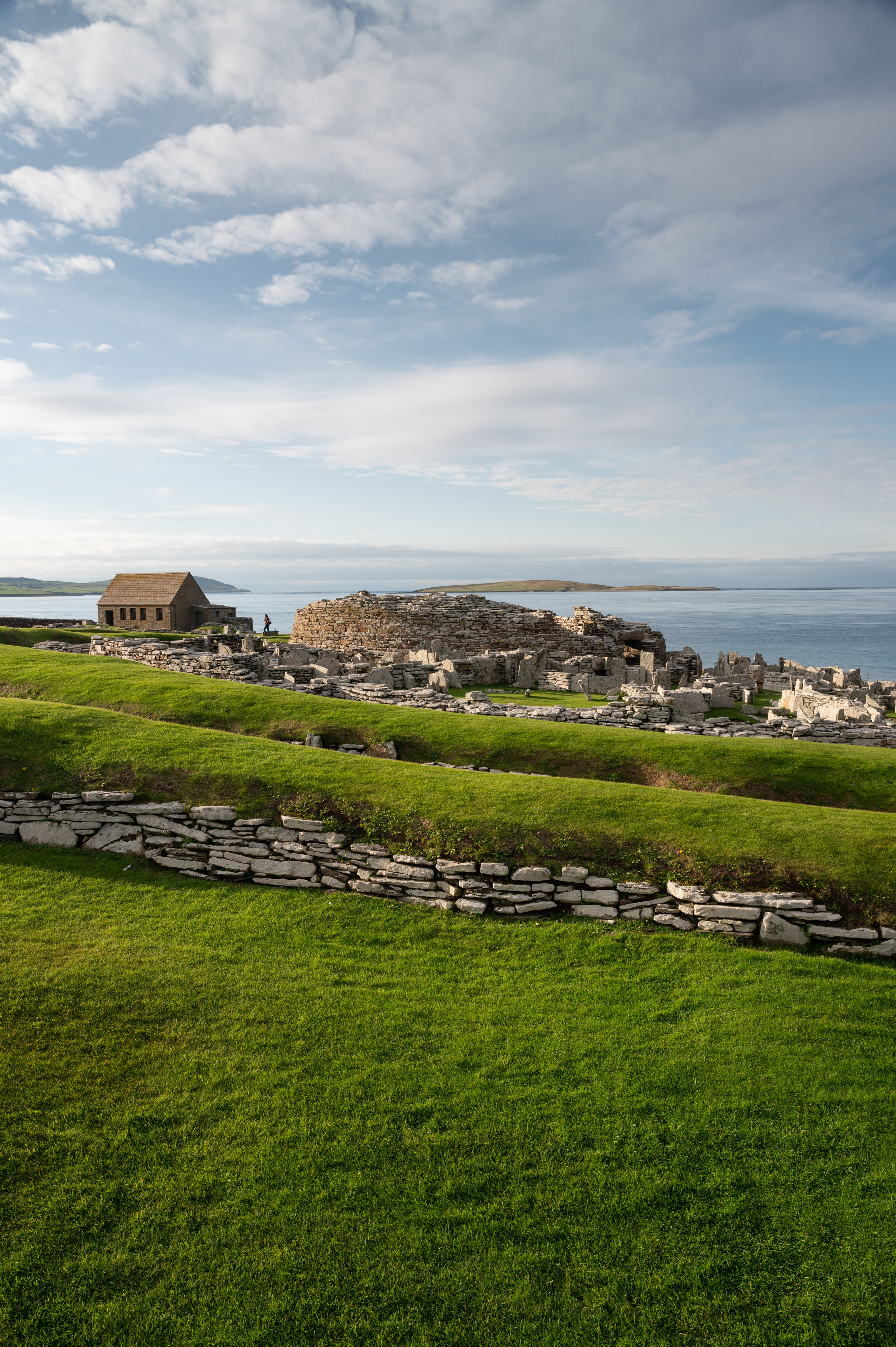 Broch ruins with green grass terraces and stone walls, overlooking a calm sea and distant islands under a bright blue sky with scattered clouds.