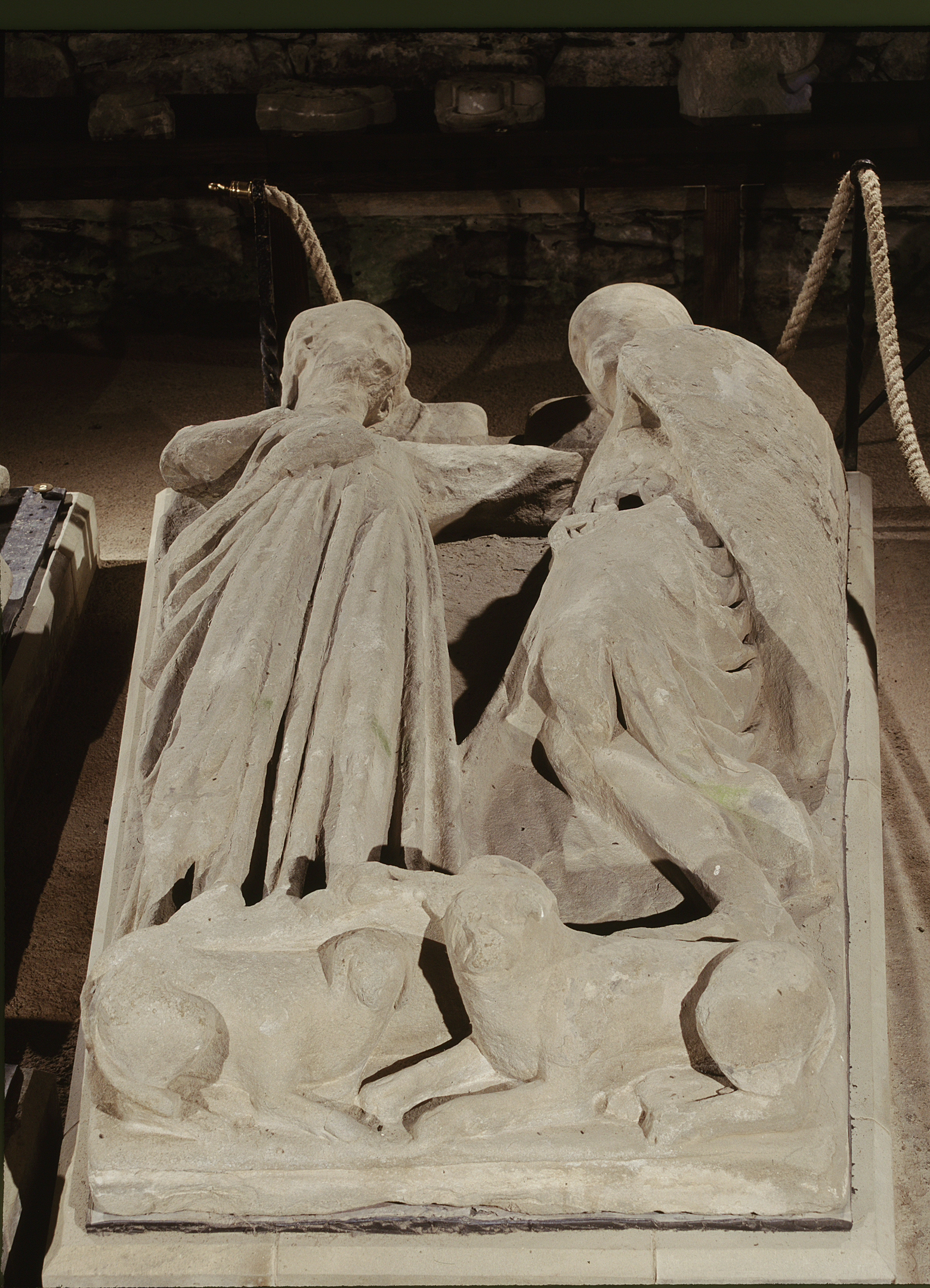 A weathered stone effigy of two reclining figures with draped robes and carved animals at their feet.