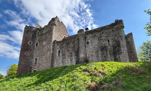 A stone castle sits on top a green grass hill. The left hand side of the castle is a tall rectangular shape with a shorter, longer section towards the right hand side. There are a number of gaps along the walls where windows once were. It is a sunny day with a bright blue sky and some clouds above the castle.