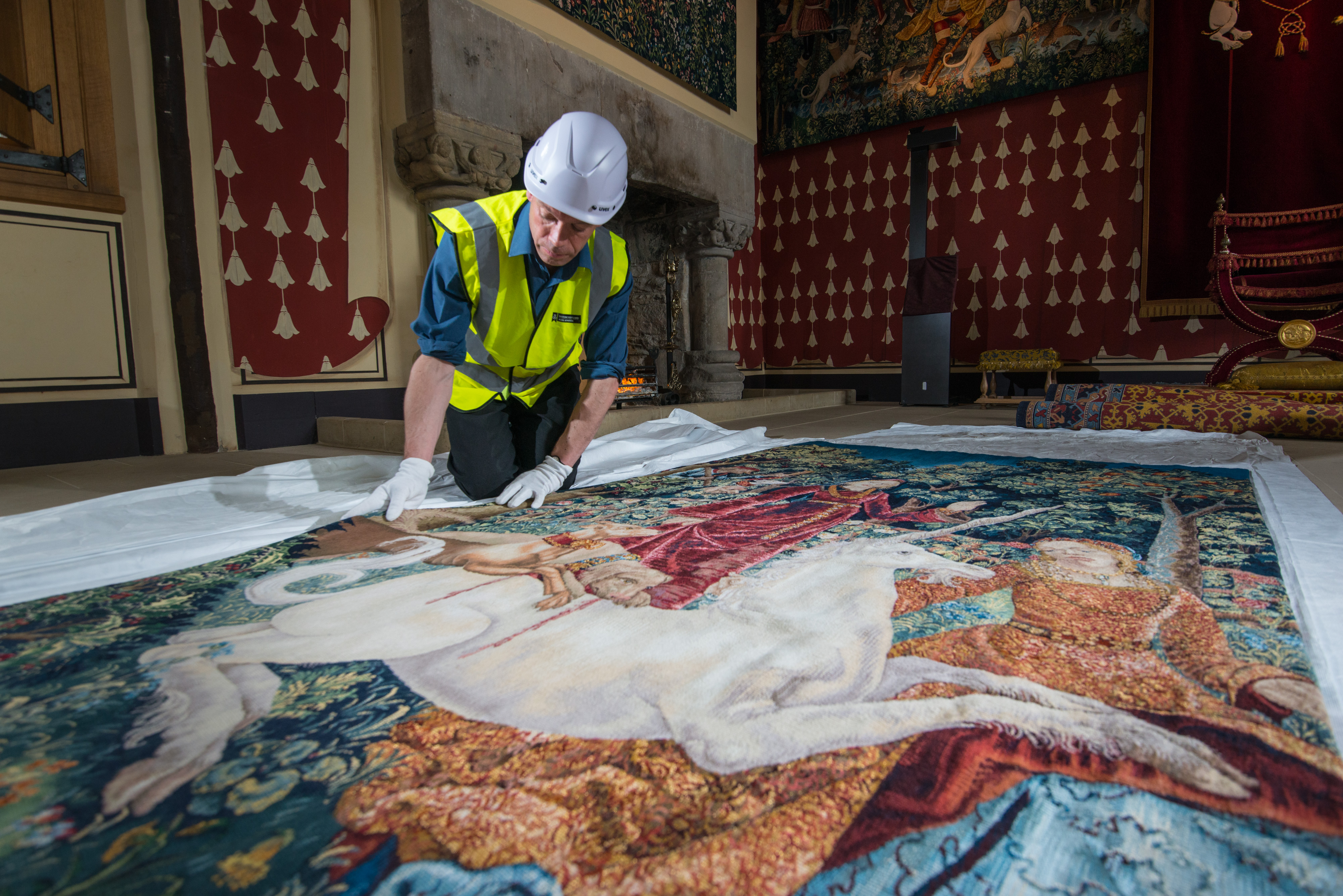 A conservator wearing a hard hat and high‑visibility vest carefully smooths a large medieval tapestry laid out on protective sheets inside a richly decorated historic hall.