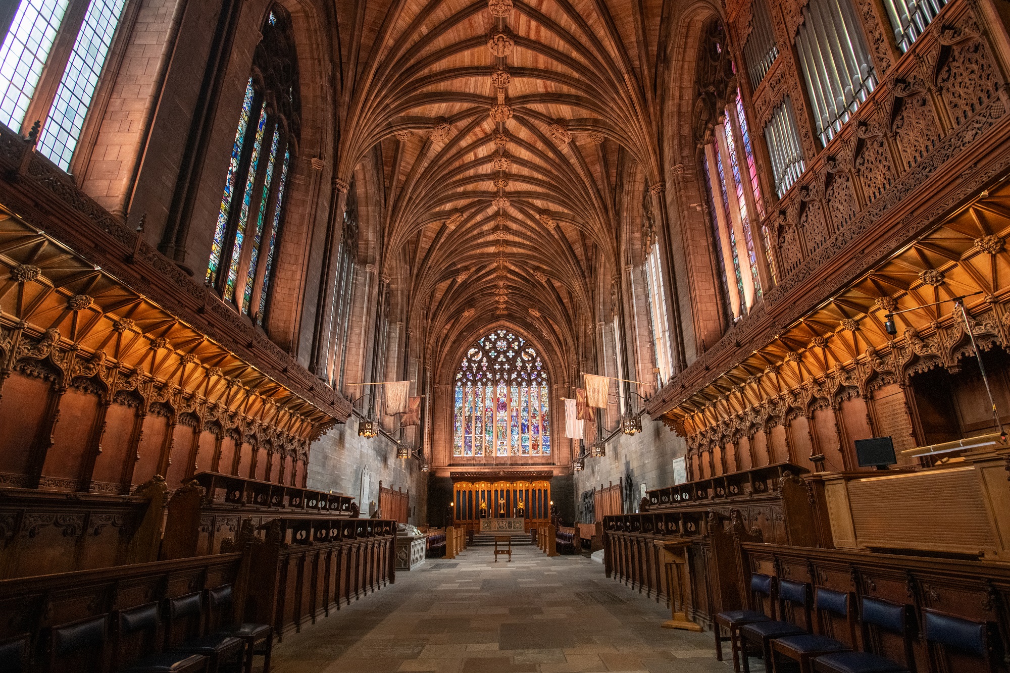 View down the choir of a large Gothic cathedral, featuring carved dark wooden choir stalls on either side, a vaulted stone ceiling with intricate ribbed arches, and tall stained glass windows. At the far end, an ornate stained glass window stands above the altar area