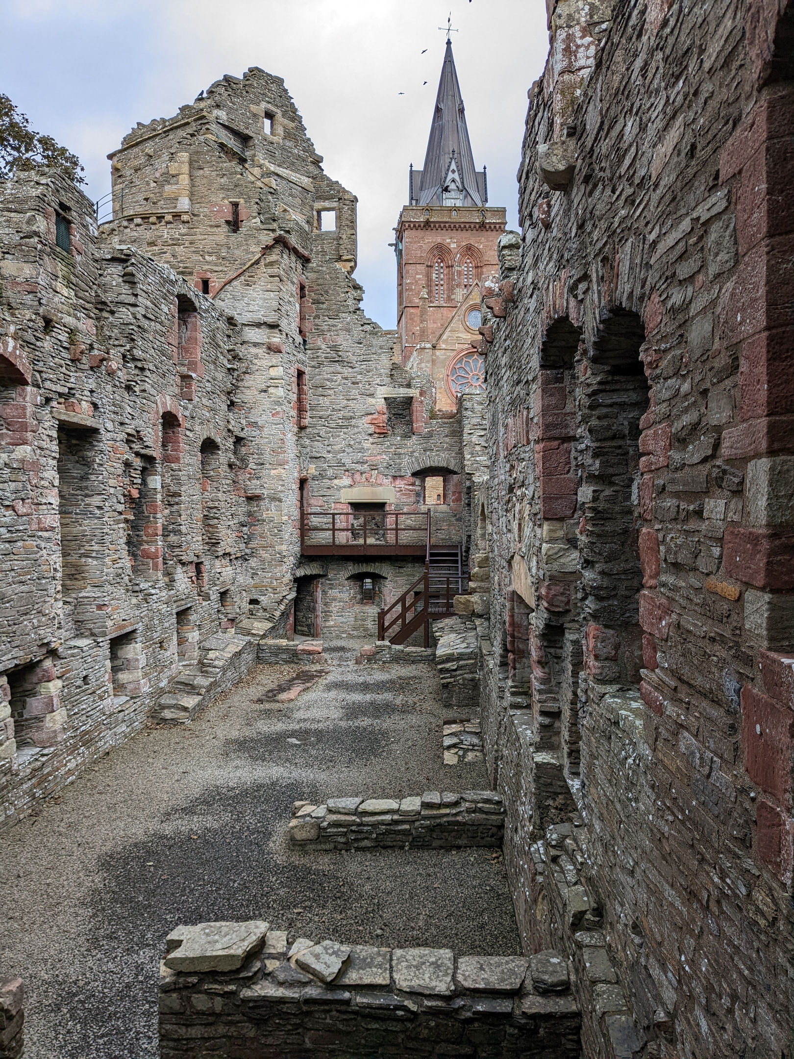 A long, narrow interior courtyard of a stone palace ruin, with tall weathered walls, arched openings, and a church spire visible above the structure in the background.