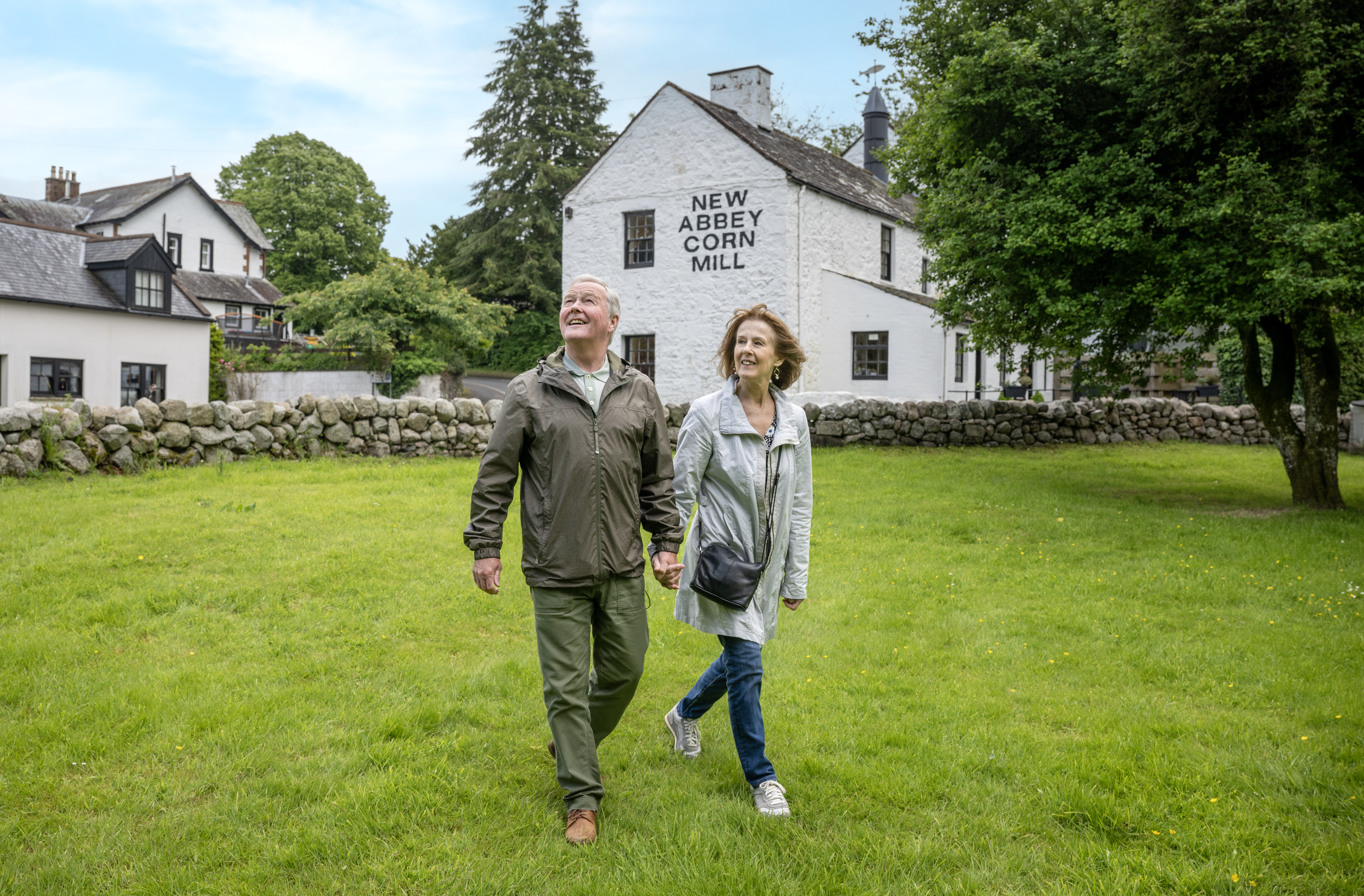 Couple smiling walking on grass. A white building with the words 'New Abbey Corn Mill' is behind them. 