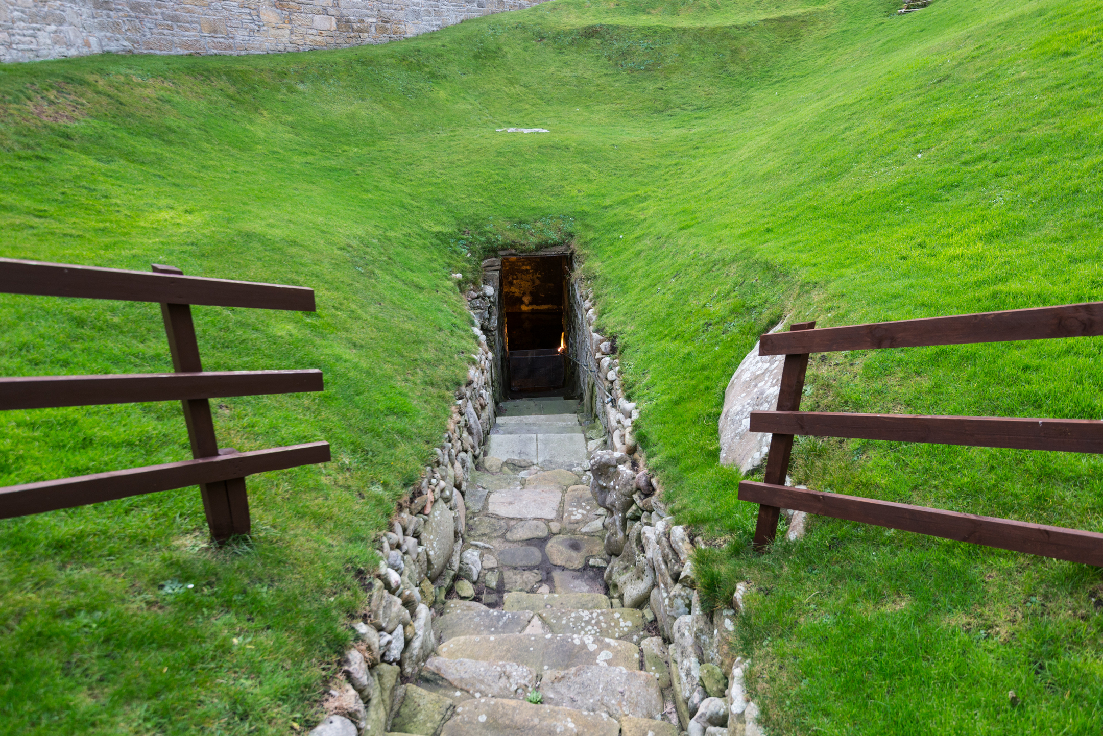 View of Burghead Well entrance from outside, showing a narrow stone-lined passage descending into the ground. The passage is flanked by grassy slopes and wooden railings, leading to a dark doorway at the bottom. The surrounding area is enclosed by stone walls.