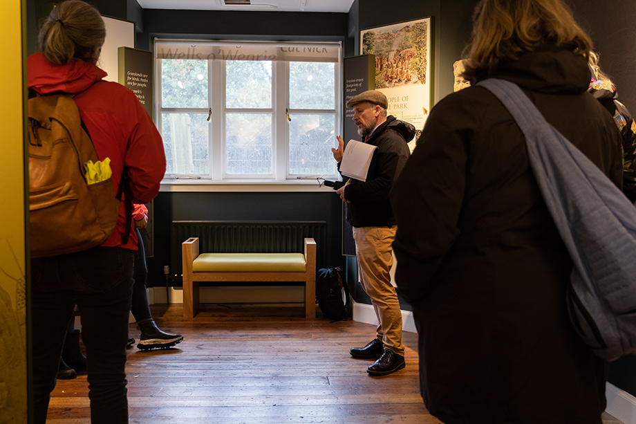 A man wearing a flatcap and holding a brolly in one hand and a flipboard under his arm talks to a group of people indoors by a set of windows. Two people wearing backpacks stand either side of the picture, facing towards the window and speaker
