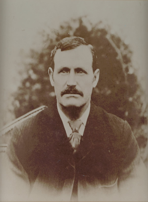 Sepia toned, front on portrait photo of gentleman in a suit, with a large moustache and short black hair parted on the side.