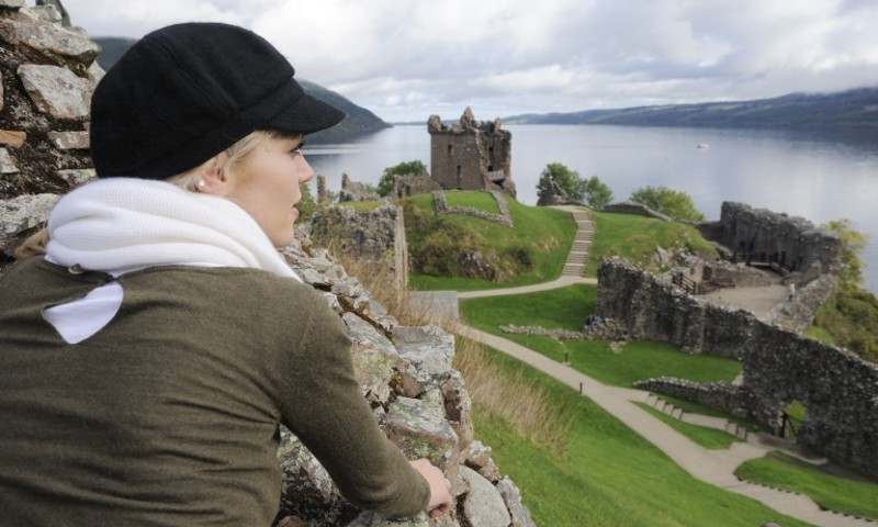 A photograph of a blonde woman in a hat look out over a castle and lake behind it