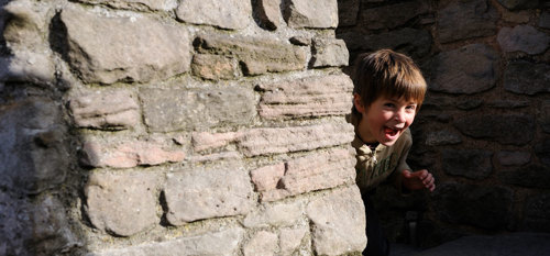 A smiling boy creeps round a corner from hiding behind a wall