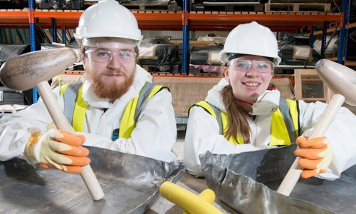 bearded man and woman with ponytail pose with lead shaping tools, both dressed in hard hats, safety googles and hi-vis vests