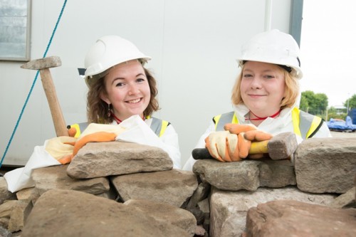 Two women in hard hats with mallets standing behind a stone wall