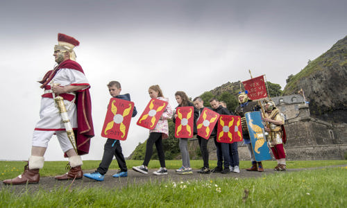 costumed performer in roman toga, cape and helmet leads parade of 6 school children holding red and gold shields with two more romans at the back 