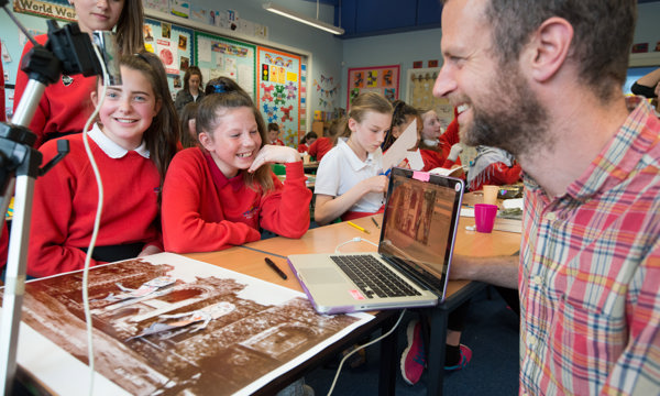 three caucasian girls in red jumpers and one in a white blouse at a table with laptop in front of them and man in plaid shirt showing them a programme on screen