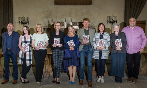 9 people stand in a row in the great hall at stirling castle, 7 holding a book with a white cover and red map of scotland with black text that reads 'bloody scotland'