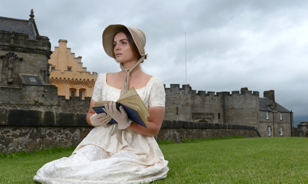 caucasian woman dressed in white dress, straw bonnet and white gloves sits on grass holding an old blue book with castle walls behind her