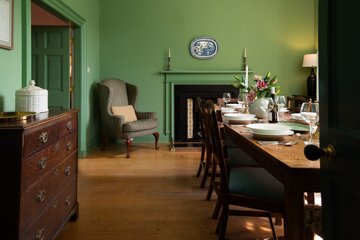 A green dining room with wooden floor, large wooden dining table set with plates and decorated with flowers, a large armchair and old, dark wooden chest of drawers