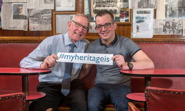 Two men with glasses sitting in a pub, holding a sign which reads #myheritageis