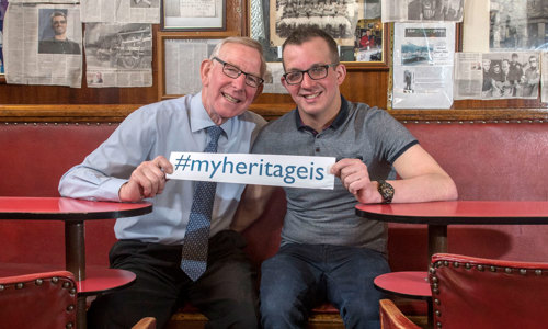 Two men with glasses sitting in a pub, holding a sign which reads #myheritageis