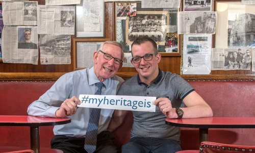 Two men with glasses sitting in a pub, holding a sign which reads #myheritageis