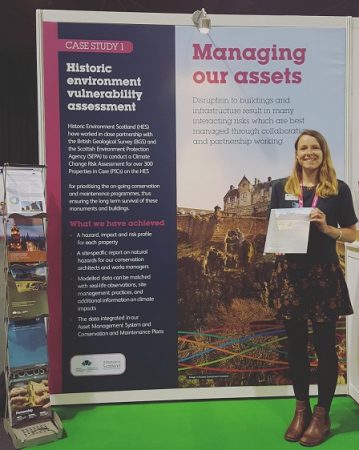 Woman stands in front of a banner that reads 'Managing our assets' with sub-heading 'Historic environment vulnerability assessment'