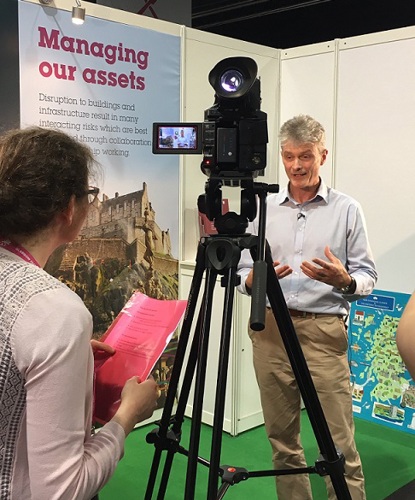 A man being interviewed by a woman holding a red document, with a video camera pointed at the man, with a banner in the background with the header 'Managing our assets'