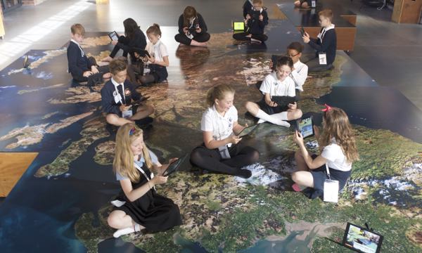 a group of school children sit on a map of Scotland holding iPads 