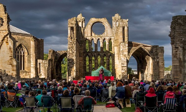 A photograph of a crowd of people watching performers on stage in the ruins of an old cathedral at sunset
