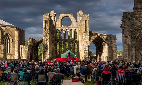 A photograph of a crowd of people watching performers on stage in the ruins of an old cathedral at sunset