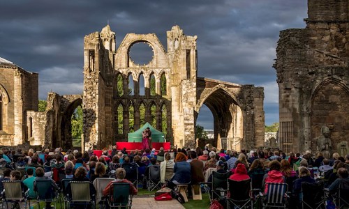 A photograph of a crowd of people watching performers on stage in the ruins of an old cathedral at sunset