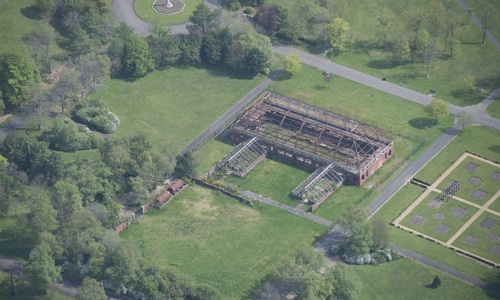 aerial view of a green space with the shell of a building with stone walls and iron supporting beams that have fallen into disrepair