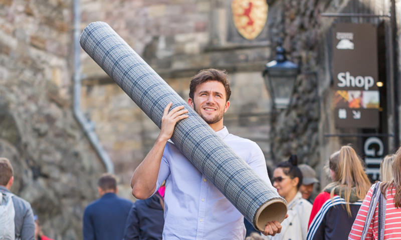 A photograph of a man in a kilt carrying a large roll of grey and blue tartan at the entrance to a castle, surrounded by tourists