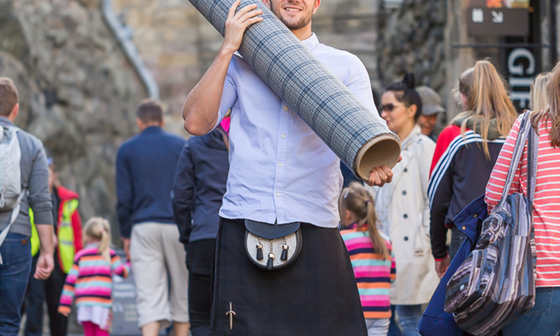 A photograph of a man in a kilt carrying a large roll of grey and blue tartan at the entrance to a castle, surrounded by tourists