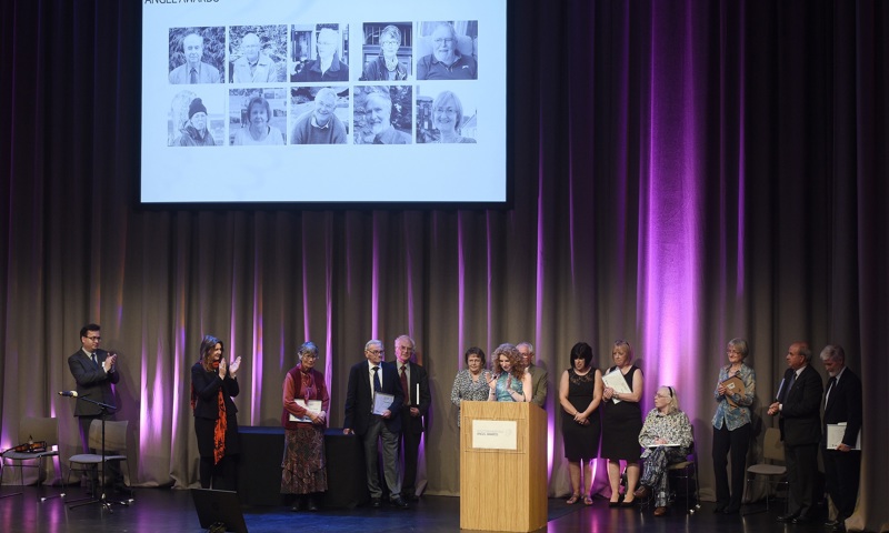 A photograph of a stage filled with people, listening to a woman speaking at a podium and applauding.