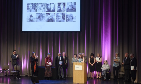 A photograph of a stage filled with people, listening to a woman speaking at a podium and applauding.