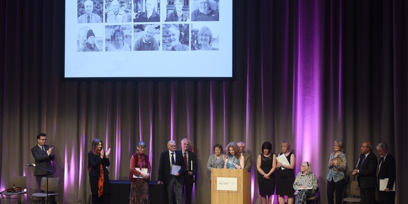 A photograph of a stage filled with people, listening to a woman speaking at a podium and applauding.