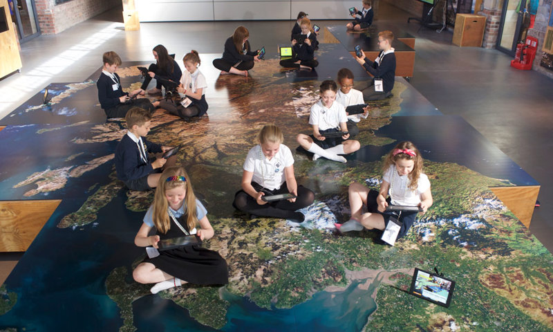 A photograph of children sitting on a large map of Scotland looking at tablets
