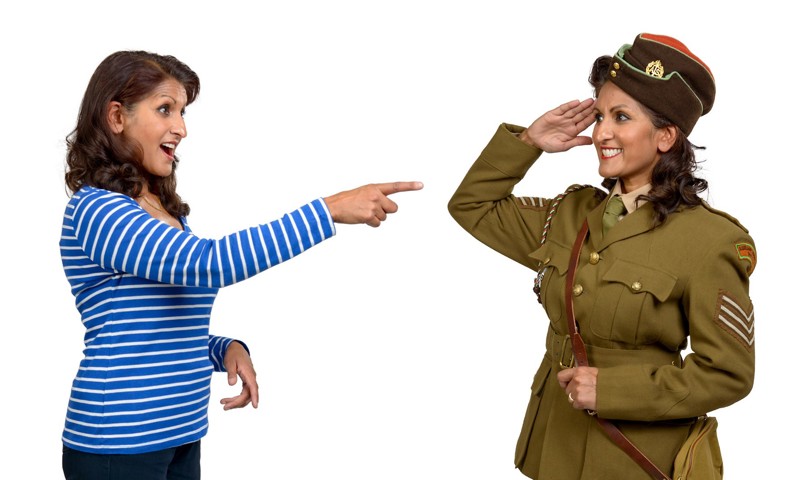 A photograph of a woman in a stripey blue top pointing in surprise at her mirror image who is dressed in a 1940s uniform, saluting nearby