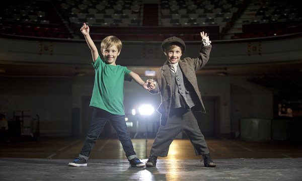 twin boys stand side by side with arms in air smiling, one dressed in jeans and t-shirt while brother is in victorian clothes