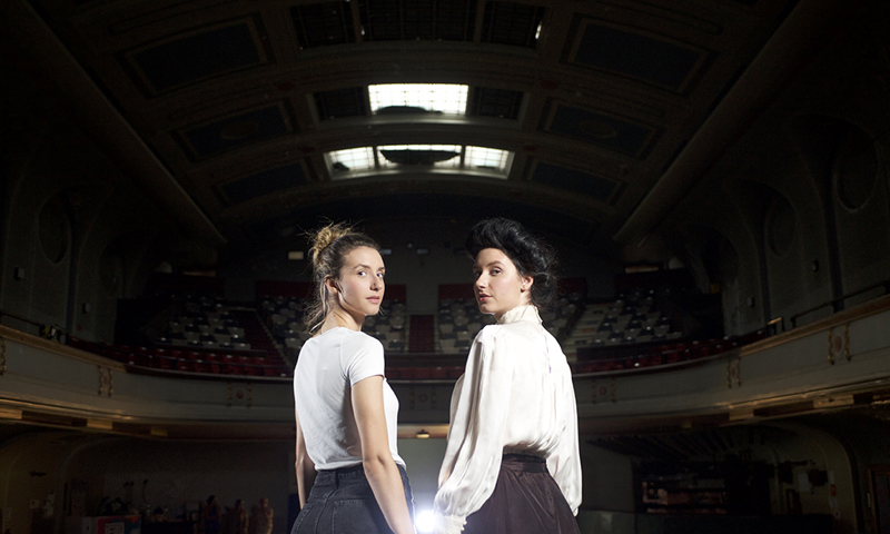 female twins stand side by side looking over shoulders one dressed in jeans and t-shirt other in victorian clothes