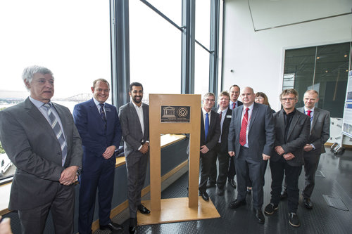 A group of business executives stand for a photo around a plaque with large windows behind them