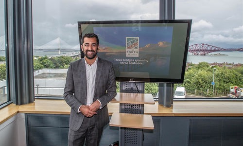 Man at Forth Bridges office standing in front of the windows overlooking the bridges, next to a TV screen