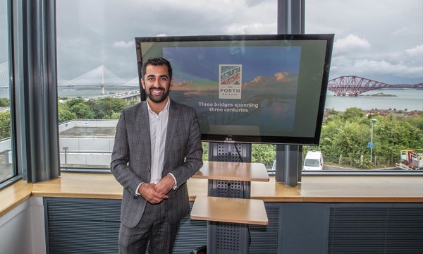Man at Forth Bridges office standing in front of the windows overlooking the bridges, next to a TV screen