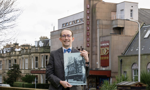 A man smiles at the camera whilst holding a large black and white photo of an art deco cinema building. He wear a grey suit, white shirt, blue striped bowtie and glasses. The cinema from the photo is in the background behind him.